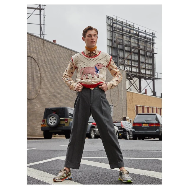 A young adult male poses on a city street wearing an animal print sweater vest, collared shirt, grey slacks, and sneakers with buildings in the background.