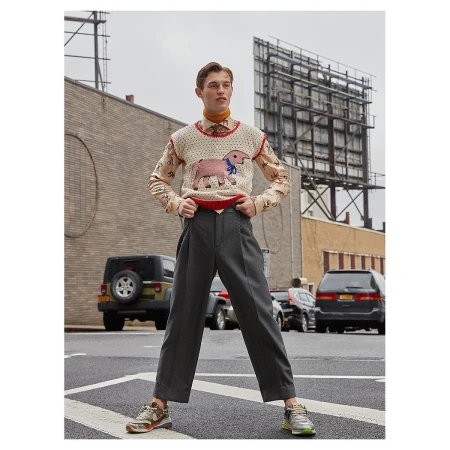 A young adult male poses on a city street wearing an animal print sweater vest, collared shirt, grey slacks, and sneakers with buildings in the background.