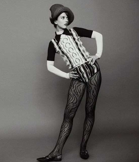 Woman in patterned knitwear, dark tights, and a straw hat poses against a gray backdrop in a full-length studio shot.