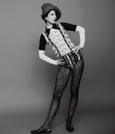 Woman in patterned knitwear, dark tights, and a straw hat poses against a gray backdrop in a full-length studio shot.