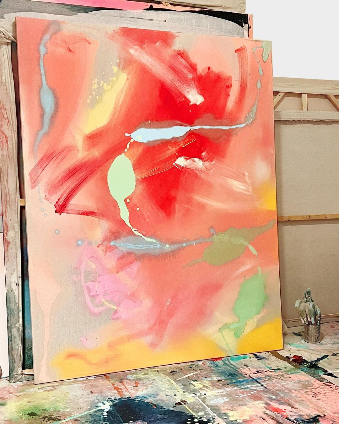 Abstract painting in orange, red, yellow, green, pink, and blue with expressive brushstrokes, shown in an artist's studio setting.