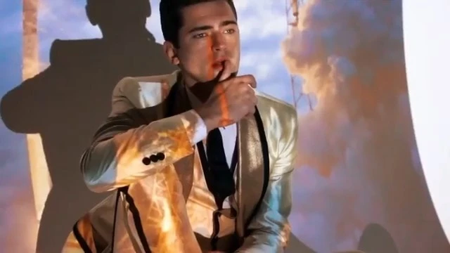 Adult male in a shiny gold suit poses in a photo studio with projected clouds and sky and a dark shadow on the left side.