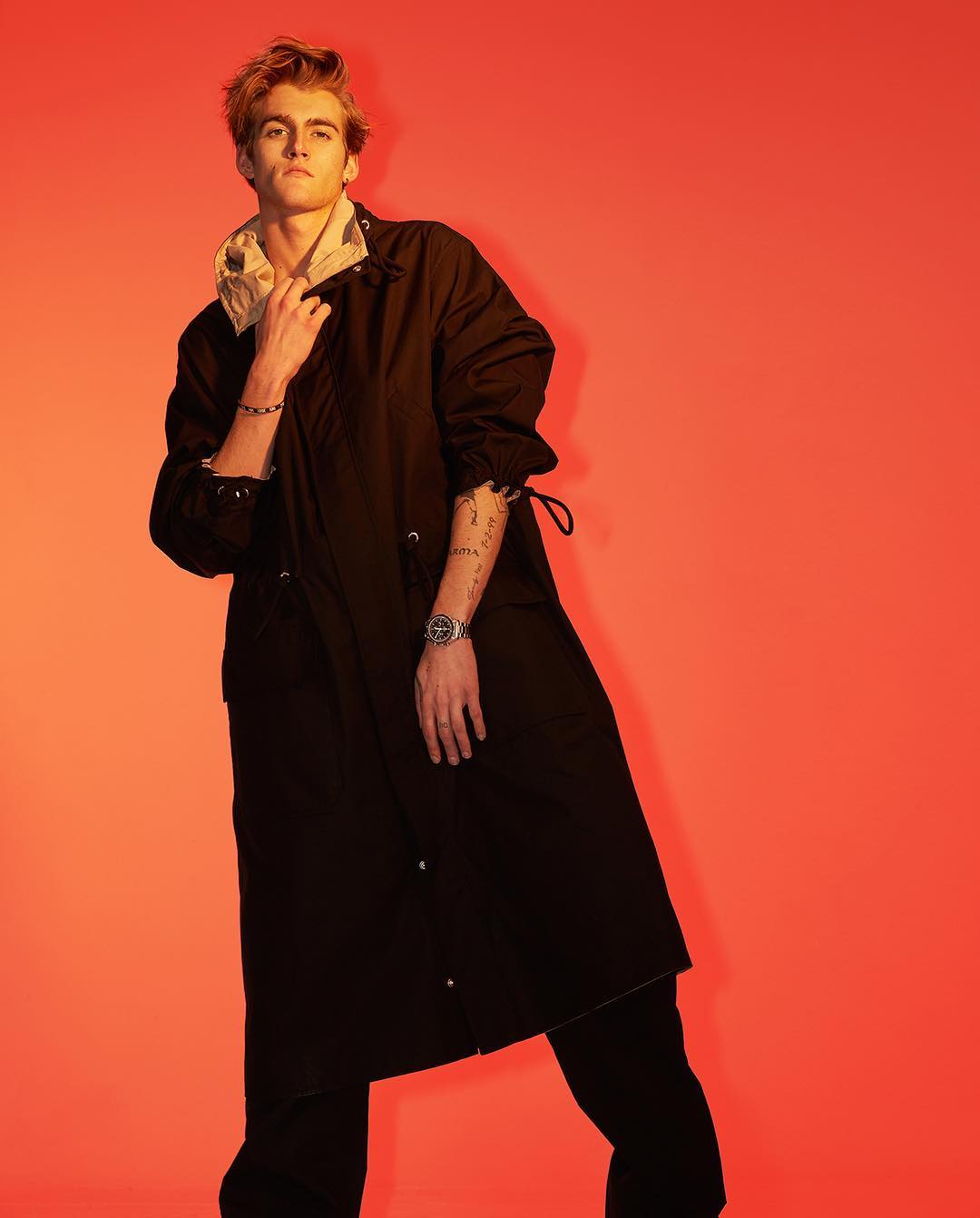 Stylish young adult male in black overcoat poses in a studio with a bright red background, showcasing fashion and portrait photography.