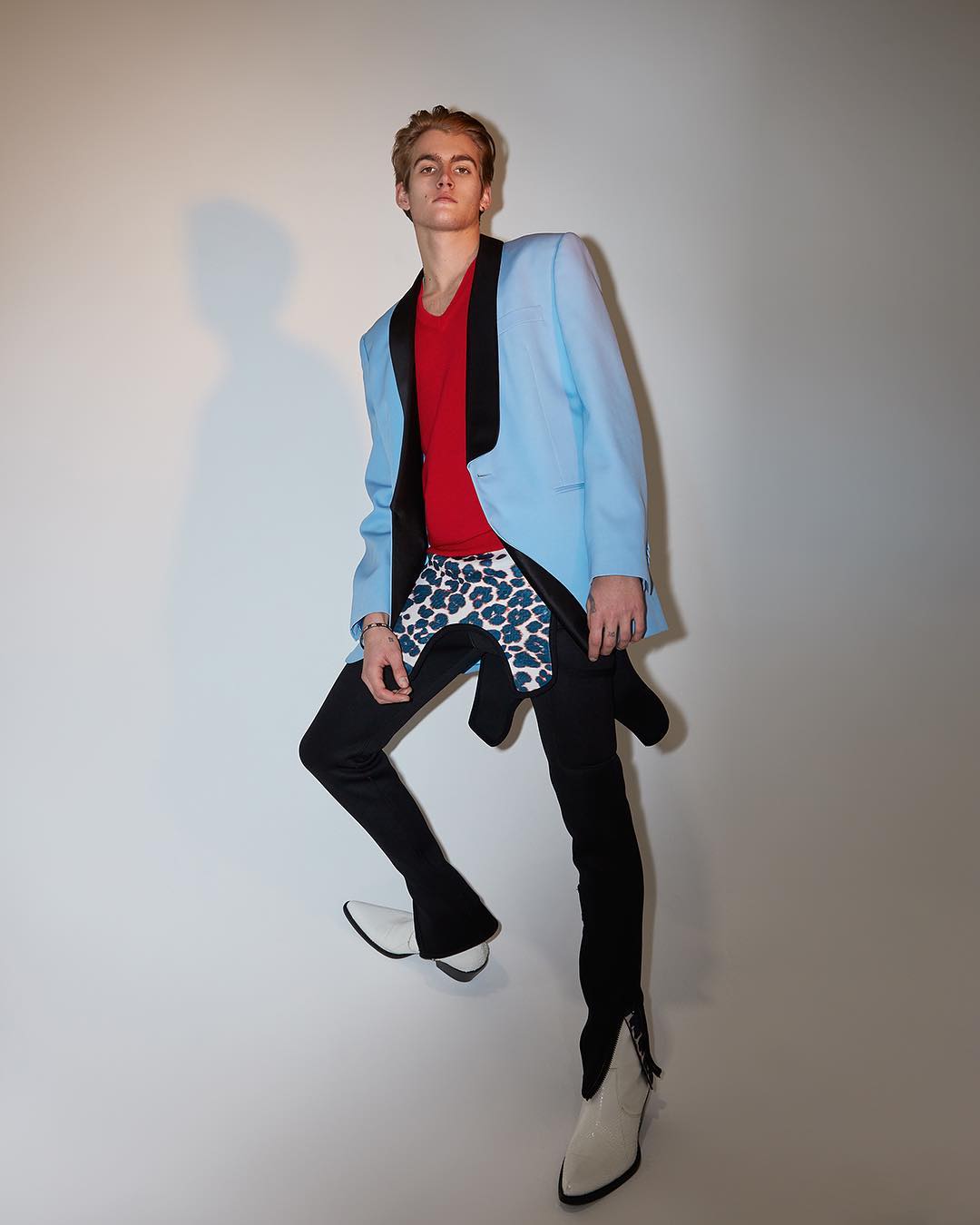 Young adult male in a light blue blazer, red shirt, patterned shorts, and white boots strikes a high-fashion pose in a photo studio.