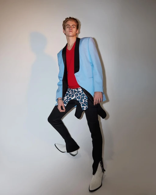Young adult male in a light blue blazer, red shirt, patterned shorts, and white boots strikes a high-fashion pose in a photo studio.