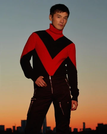 Portrait of a man in black and red sweater and black pants with zippers standing against a sunset sky and distant cityscape.