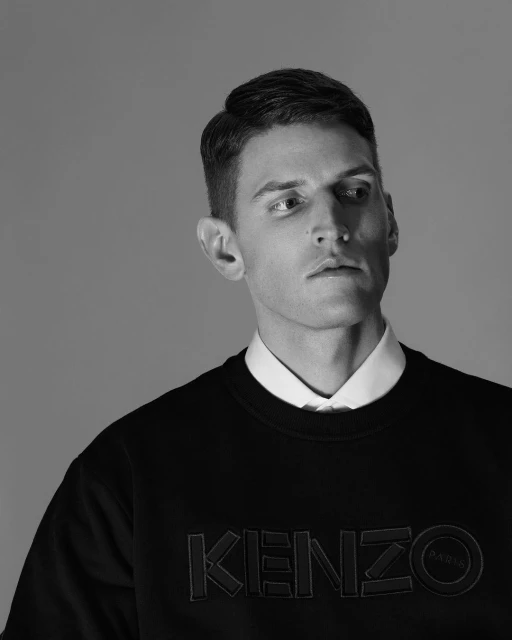 Monochrome portrait of a handsome man in a black KENZO sweatshirt and collared shirt against a grey backdrop with studio lighting.