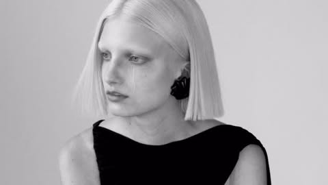 Monochromatic portrait of a platinum blonde adult female model wearing black dress and geometric earrings against a plain white backdrop.