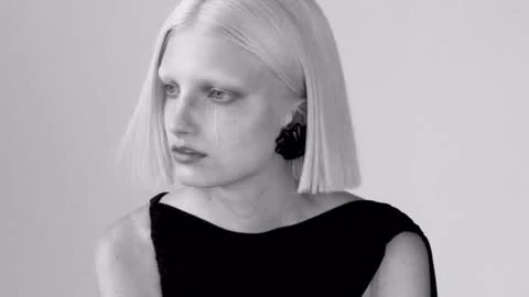 Monochromatic portrait of a platinum blonde adult female model wearing black dress and geometric earrings against a plain white backdrop.