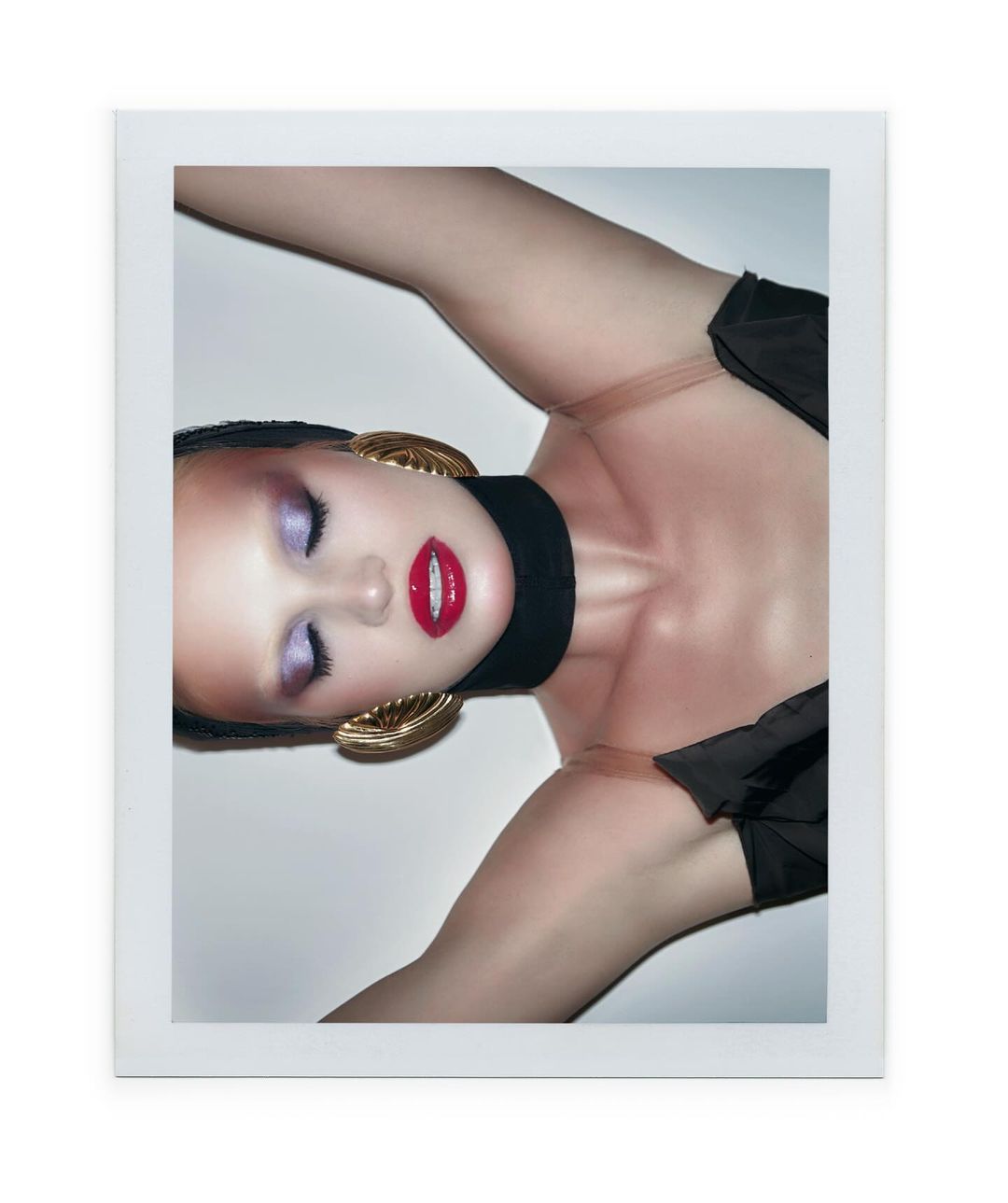 Beauty shot of a woman lying down, wearing gold earrings and dark makeup with bright red lipstick, against a white background.