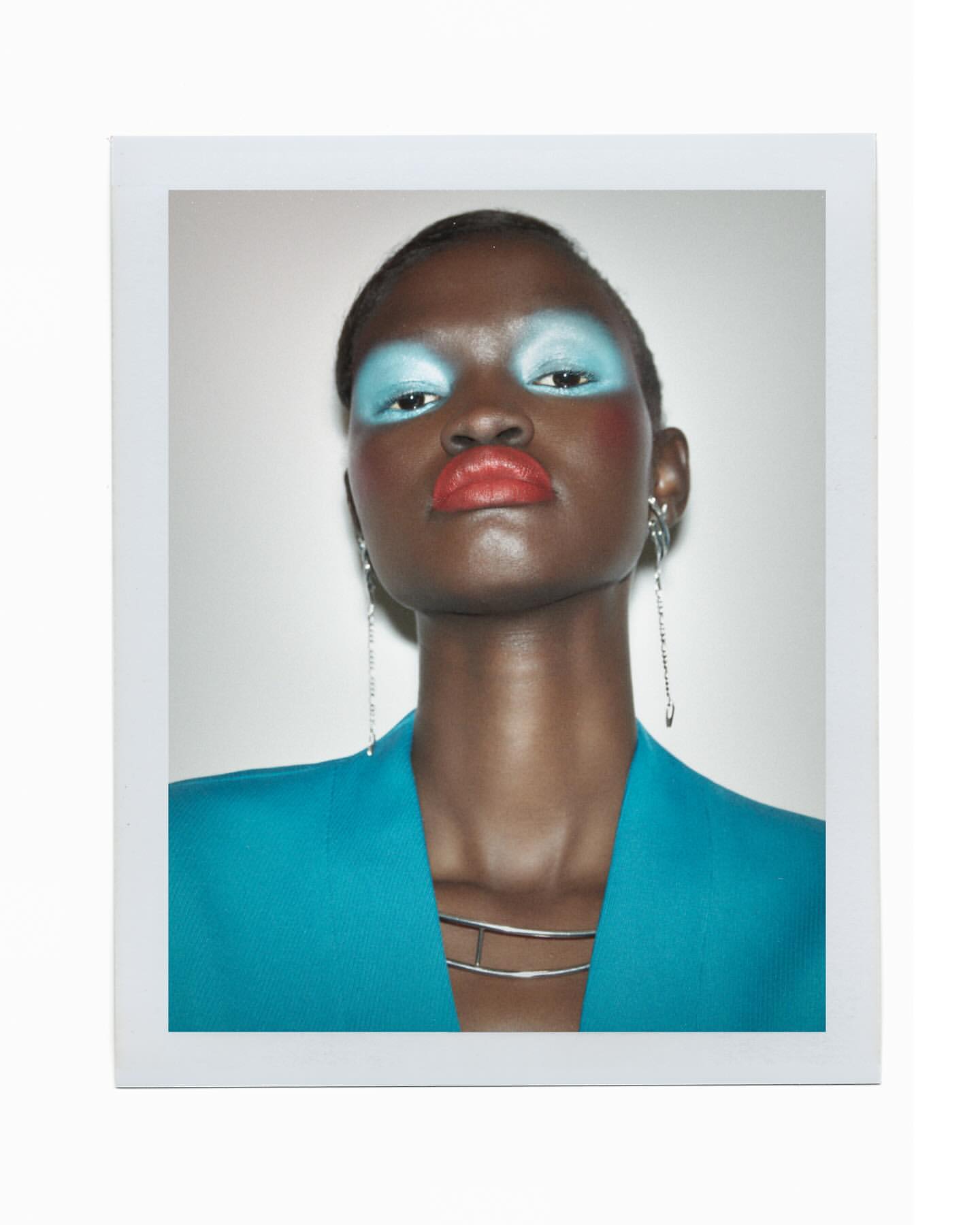 Close-up portrait of a Black woman with blue eyeshadow and coral lipstick wearing a teal blazer against a neutral backdrop.