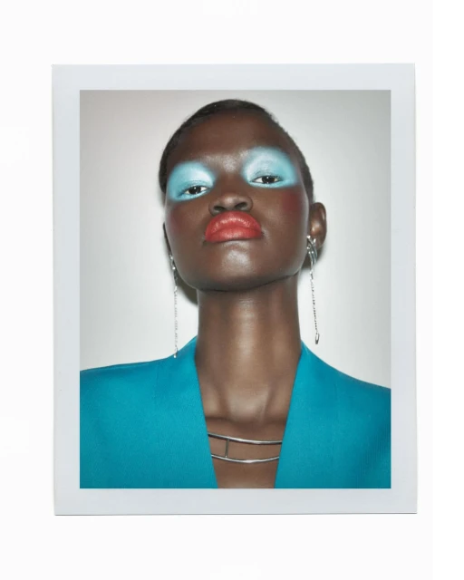 Close-up portrait of a Black woman with blue eyeshadow and coral lipstick wearing a teal blazer against a neutral backdrop.