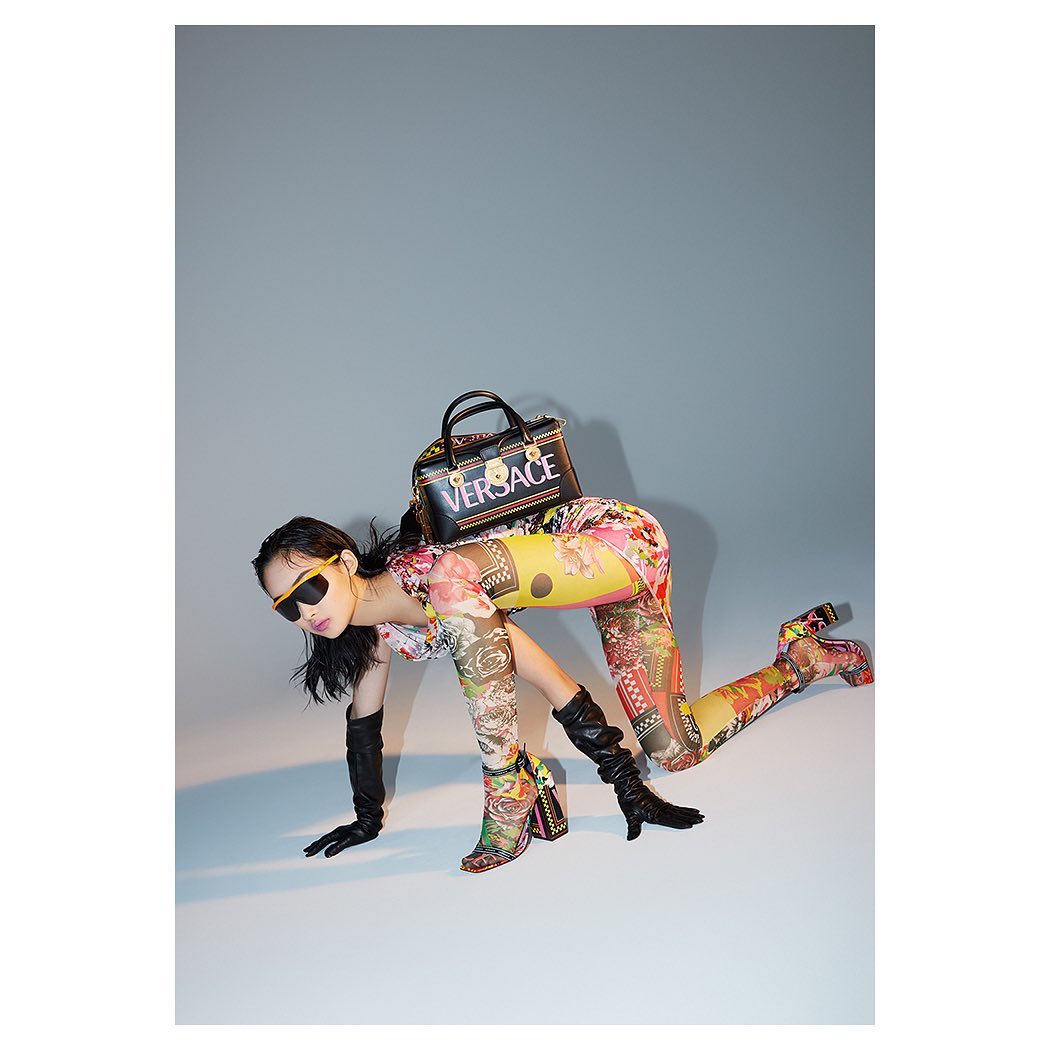 Woman in floral bodysuit poses in studio with Versace purse and boots. The lighting is bright with a neutral grey background.