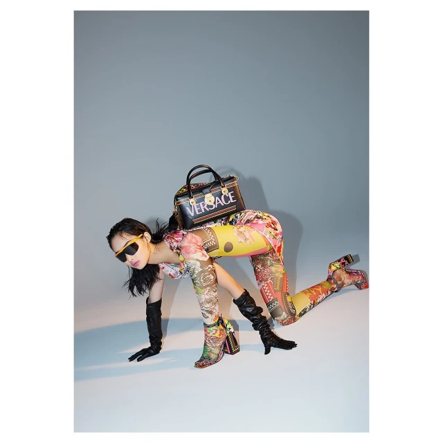 Woman in floral bodysuit poses in studio with Versace purse and boots. The lighting is bright with a neutral grey background.