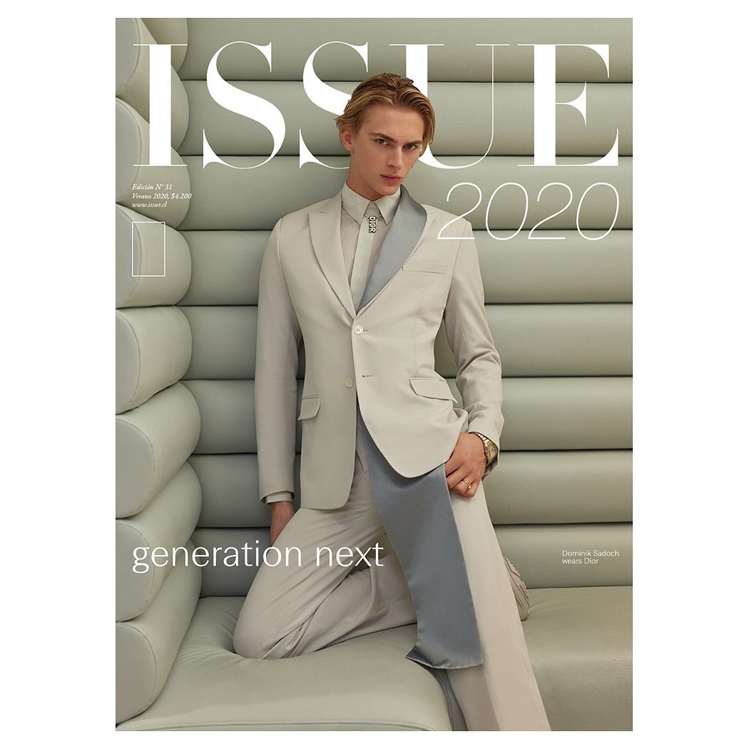 Fashion magazine cover featuring a blond male model in a tailored beige suit with a grey lapel kneeling against a textured wall.