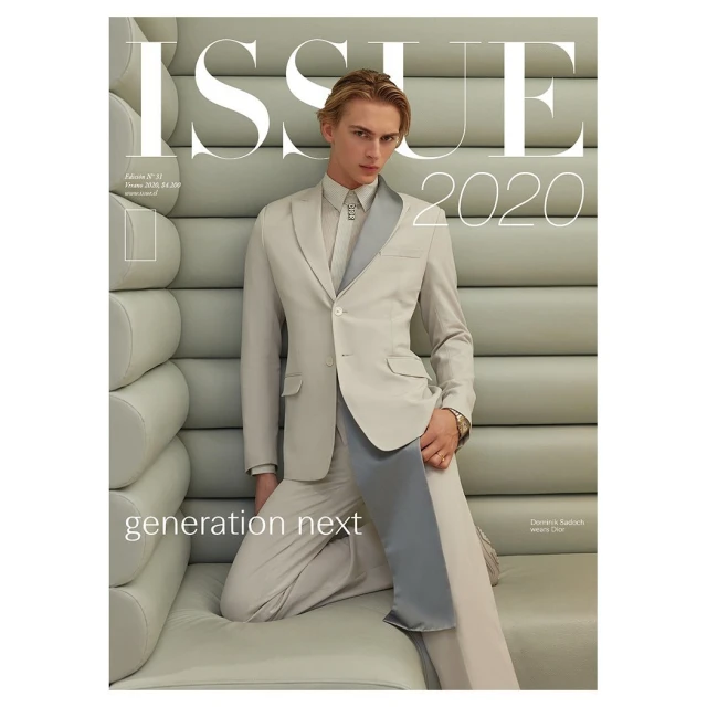Fashion magazine cover featuring a blond male model in a tailored beige suit with a grey lapel kneeling against a textured wall.