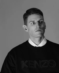 Portrait of a man in a black sweatshirt with Kenzo branding over a white collar against a plain grey background