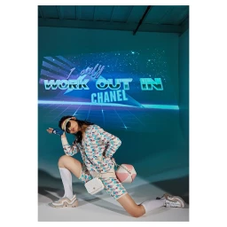 Model in patterned athletic wear kneeling on white floor with a neon projected projection background reading work out in Chanel