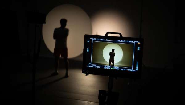 Person in silhouette poses in front of a monitor in a dimly lit studio with spotlights. The monitor displays a similar figure.