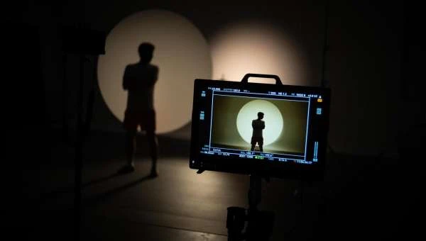 Person in silhouette poses in front of a monitor in a dimly lit studio with spotlights. The monitor displays a similar figure.