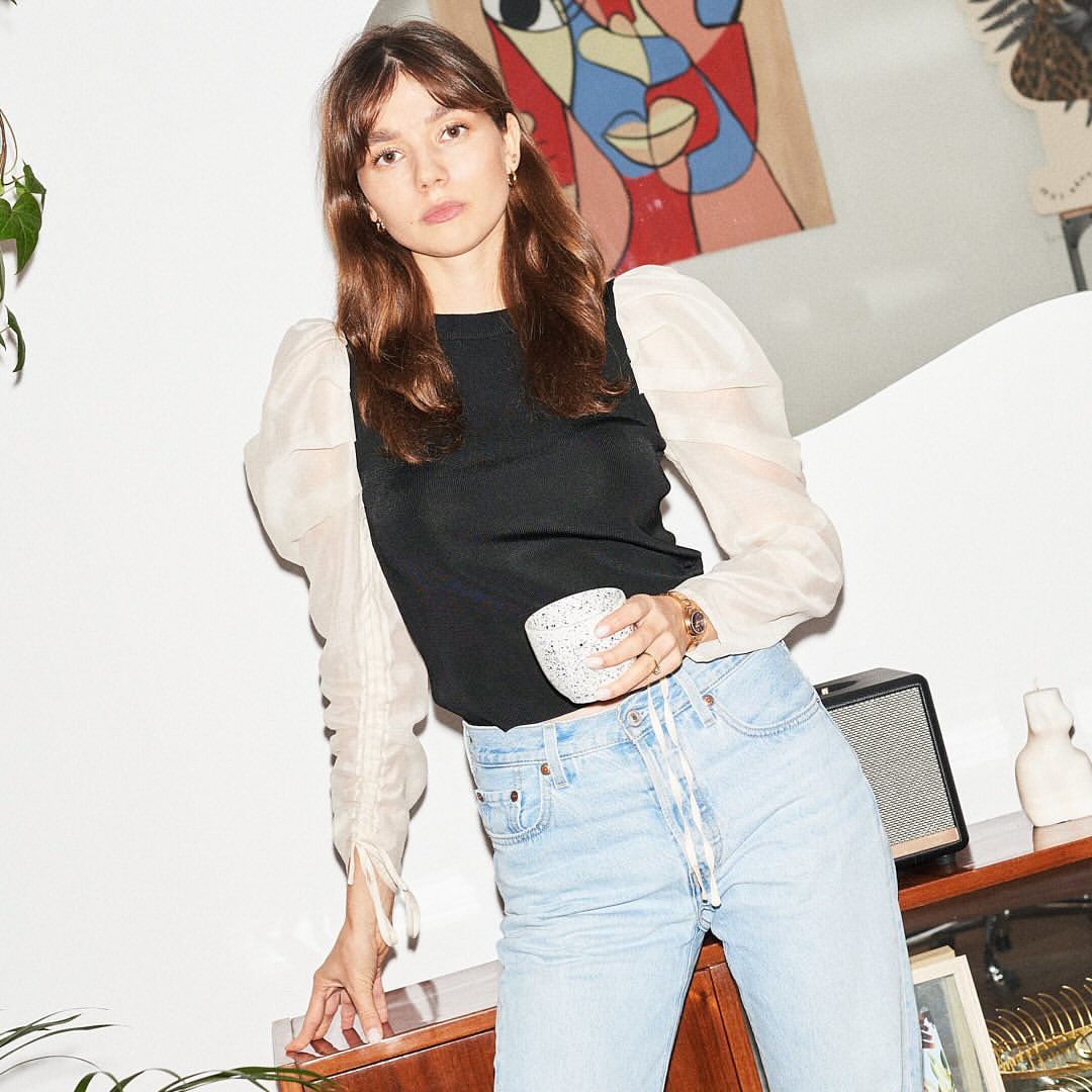 Indoor portrait of a young woman wearing a black top with sheer sleeves, holding a speckled mug, and standing near a wooden cabinet.