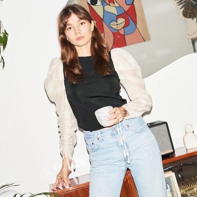 Indoor portrait of a young woman wearing a black top with sheer sleeves, holding a speckled mug, and standing near a wooden cabinet.