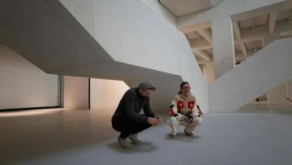 Two adults squat in a white, open space, with architectural elements and gray flooring dominating the scene. One person wears a grey hat.