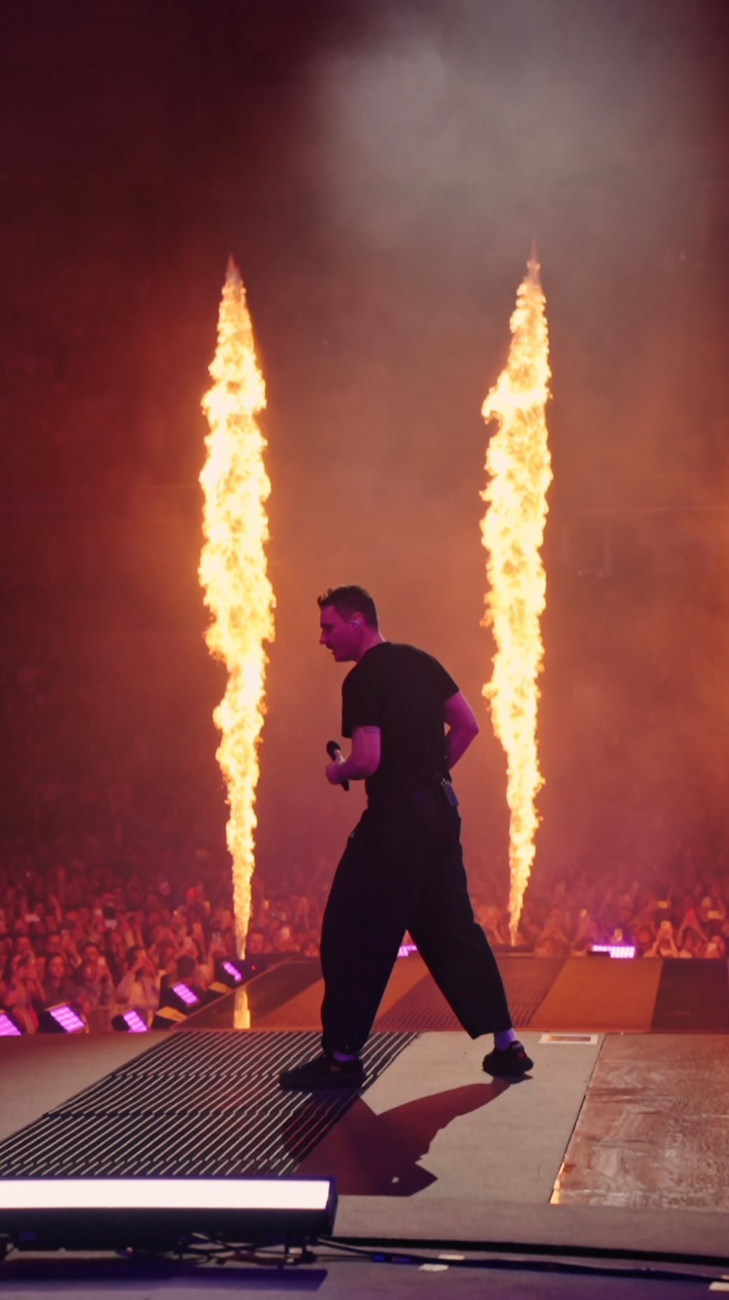 A male singer performs on stage with a microphone, wearing all black. Pyrotechnics create pillars of fire behind him as he sings.