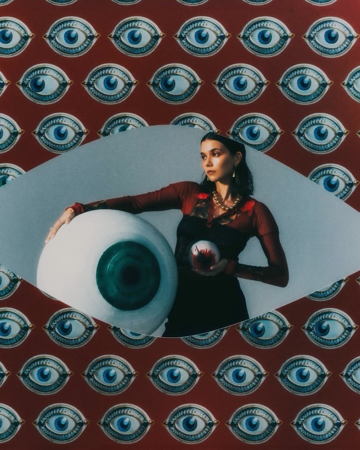 An adult woman poses with an oversized eyeball and a small orb against a backdrop patterned with painted eyes.