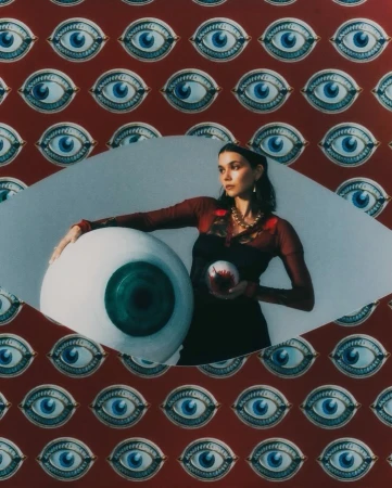 An adult woman poses with an oversized eyeball and a small orb against a backdrop patterned with painted eyes.