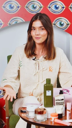 Brunette woman in a cream-colored shirt sits indoors with an assortment of skincare products on a small table in front of her.