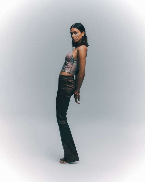 Adult female in brown pants and halter top posing in studio with silver glitter makeup around her eyes in a full shot.