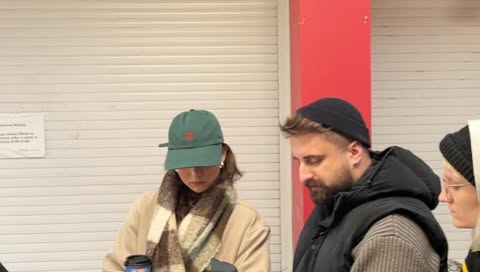 Three adults standing indoors wearing hats and jackets, one holding a coffee cup. They stand near a closed gray metal door.