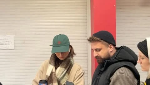 Three adults standing indoors wearing hats and jackets, one holding a coffee cup. They stand near a closed gray metal door.