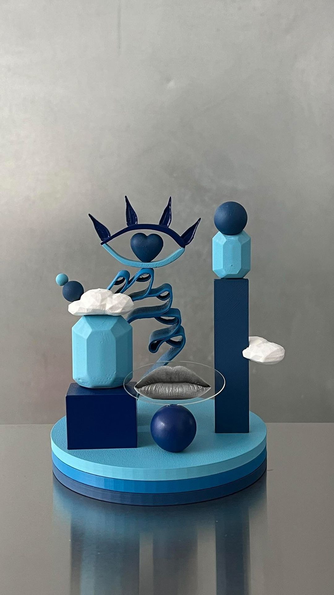 Abstract art still life features geometric shapes, eye design, lips on glass, and ball on striped platform against a silver background.