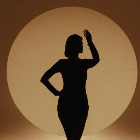 Silhouette of an adult woman in a dress posing with one hand on hip against a spotlight-lit background, creating an artistic effect.