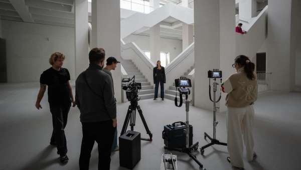 Film crew setting up camera equipment to film woman on a staircase inside a white building with large windows for natural light.