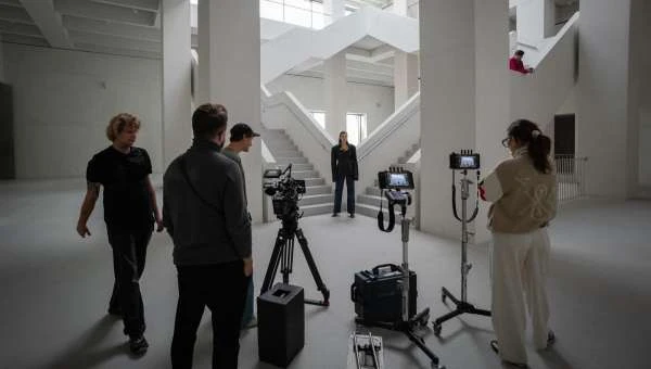 Film crew setting up camera equipment to film woman on a staircase inside a white building with large windows for natural light.