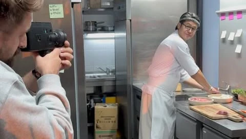 Behind-the-scenes shot of a chef in a commercial kitchen; the videographer is holding a Super 8 camera capturing the culinary scene.