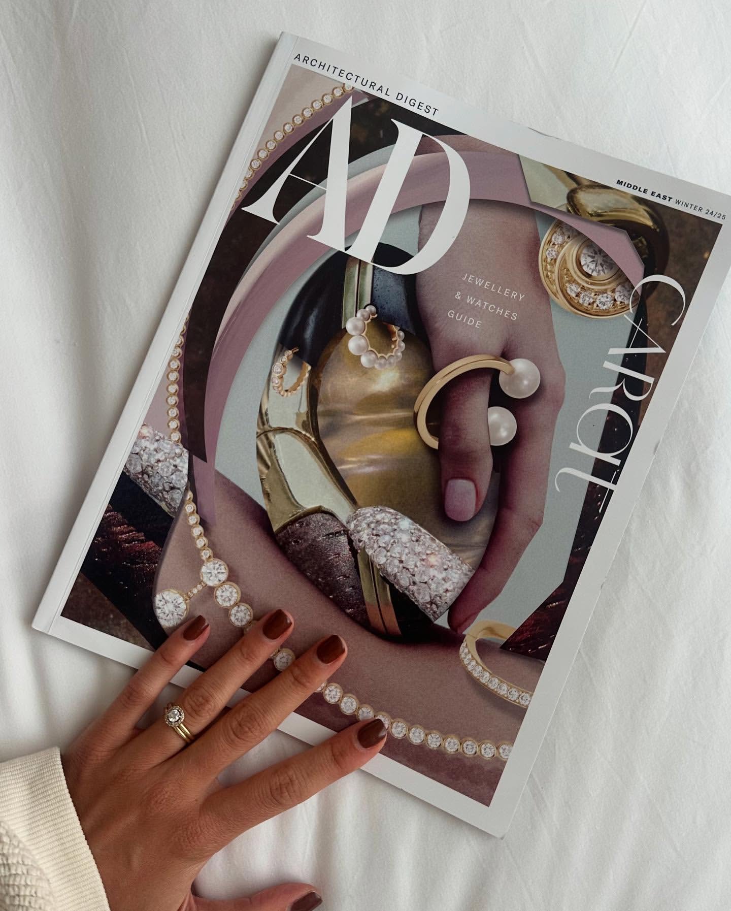 Architectural Digest magazine cover featuring jewelry, being held by a manicured hand with a ring, against a white fabric background.