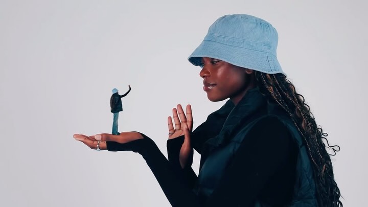 Young African woman in denim bucket hat holds miniature figure in hand against a white backdrop, creating a surreal and playful visual.