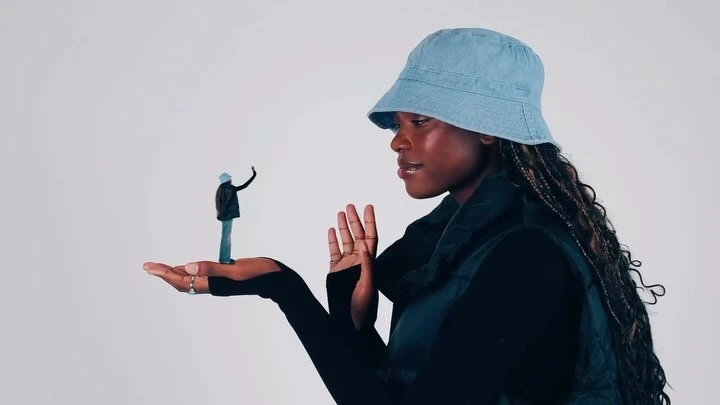 Young African woman in denim bucket hat holds miniature figure in hand against a white backdrop, creating a surreal and playful visual.