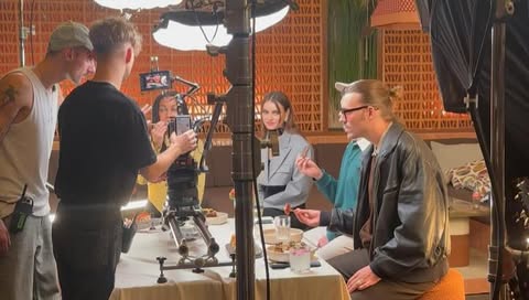 Film set with models and crew around a table with food, lights and cameras. Indoor scene with light-brown color scheme.