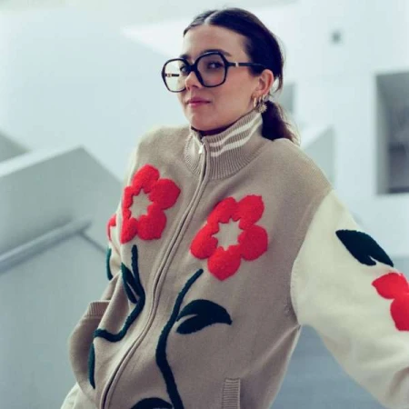 Woman in a beige zip-up flower-applique jacket and glasses stands in front of a white staircase in a medium indoor shot.