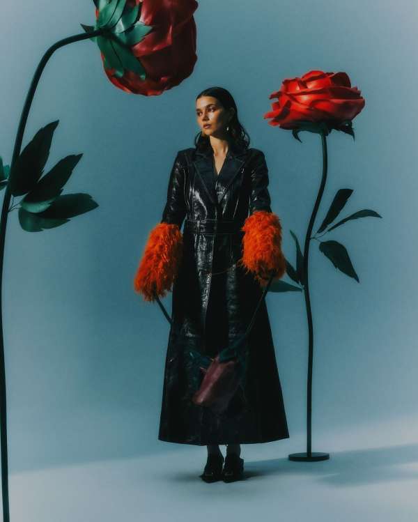 A woman in a long black coat with orange furry sleeves, standing between large red roses against a blue background in a studio shot.