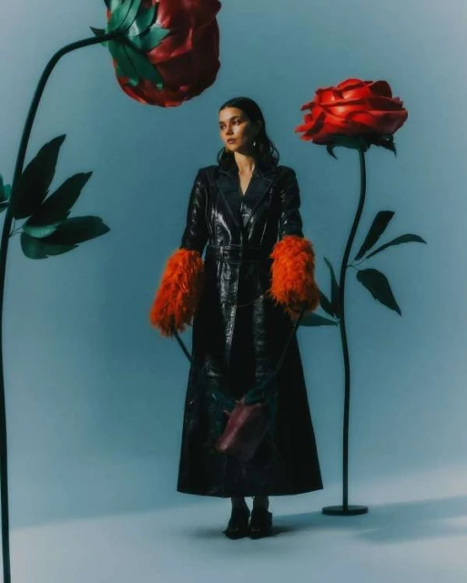 A woman in a long black coat with orange furry sleeves, standing between large red roses against a blue background in a studio shot.