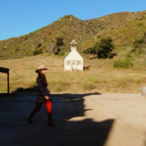 Woman in cowboy hat walks on sunny day near a white building in a rural environment with a mountain backdrop and blue sky.