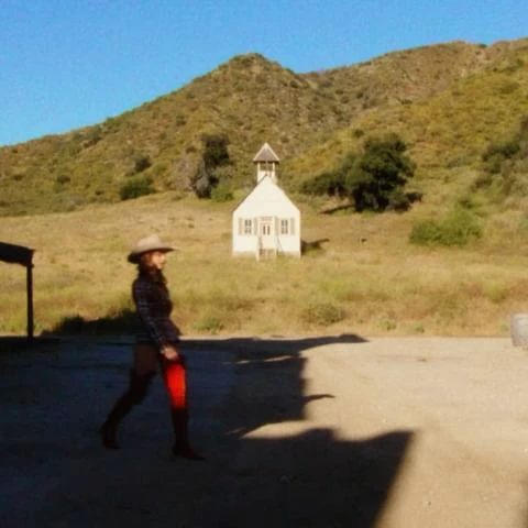 Woman in cowboy hat walks on sunny day near a white building in a rural environment with a mountain backdrop and blue sky.