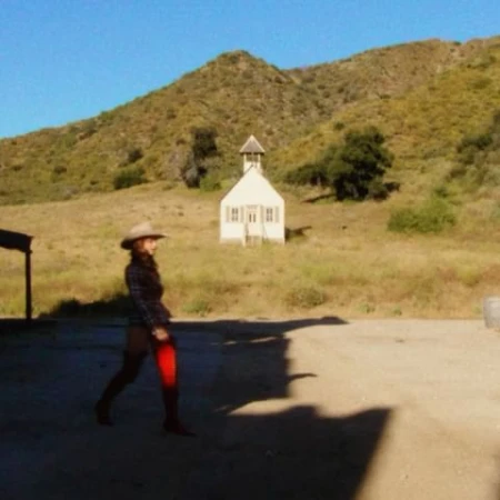 Woman in cowboy hat walks on sunny day near a white building in a rural environment with a mountain backdrop and blue sky.