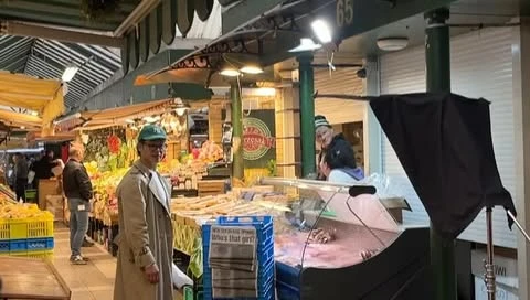 Outdoor market scene with a person in a trench coat standing near produce, a man in the background, and camera equipment visible.